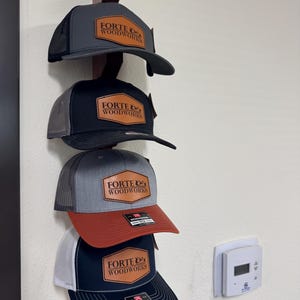 May include: Four baseball caps with the "FORTE & WOODWORKS" logo are displayed on a wooden rack. The caps feature a variety of colors, including gray, black, red, and navy blue, with mesh backs and curved brims. Each cap has a leather patch with the logo.