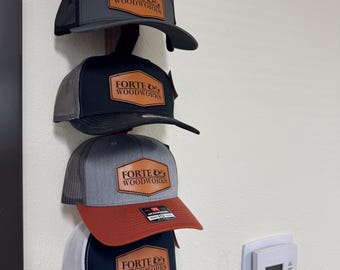 May include: Four baseball caps with the "FORTE & WOODWORKS" logo are displayed on a wooden rack. The caps feature a variety of colors, including gray, black, red, and navy blue, with mesh backs and curved brims. Each cap has a leather patch with the logo.