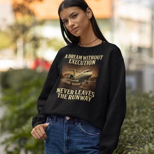 Może przedstawiać: Czarna bluza z napisem "A DREAM WITHOUT EXECUTION NEVER LEAVES THE RUNWAY" i obrazem prywatnego odrzutowca. Modelka ma na sobie niebieskie dżinsy.
