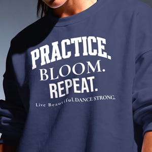 May include: A navy blue crewneck sweatshirt featuring white screen-printed text that reads PRACTICE. BLOOM. REPEAT. followed by the smaller text Live Beautiful. DANCE STRONG. The garment has long sleeves and a ribbed collar, cuffs, and hem.