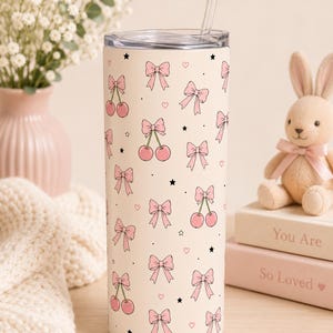 May include: A cream-coloured cylindrical tumbler featuring a repeating pattern of pink bows, pink cherries, small black stars, and tiny pink hearts. The tumbler includes a clear lid and a clear straw, providing a functional drinkware container.
