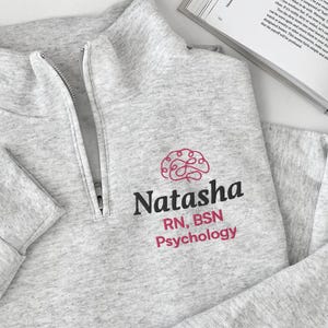 Puede incluir: Una sudadera gris jaspeado con cremallera de cuarto de largo que presenta un icono de cerebro bordado en rosa sobre el texto negro Natasha y el texto rosa RN, BSN Psychology. La prenda tiene mangas largas con puños acanalados y una cremallera plateada.