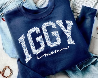 Könnte beinhalten: Marineblaues Sweatshirt mit dem Namen "IGGY" in weißer floraler Schrift und dem Wort "Mom" in Schreibschrift. Das Sweatshirt liegt auf einer Jeans.