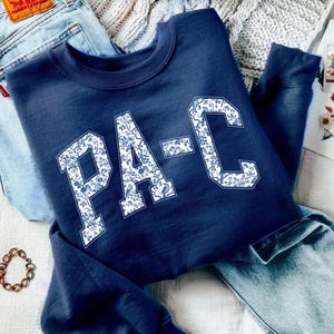 Puede incluir: Sudadera azul marino con las letras "PA-C" en blanco, con un diseño floral. La sudadera está colocada sobre unos vaqueros azul claro. La palabra "NAVY" aparece en un rectángulo azul marino en la esquina inferior derecha.