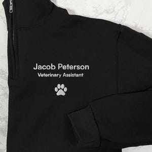 Puede incluir: Sudadera negra con cremallera de cuarto de cuello con el nombre "Jacob Peterson", el título "Asistente veterinario" y una huella de pata blanca bordada. La sudadera tiene un cuello alto.