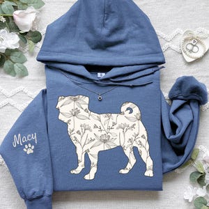 May include: A blue hoodie featuring a floral pug design. The pug silhouette is filled with a cream-colored floral pattern. The name "Macy" is printed on the sleeve, along with a paw print. A silver necklace is visible.
