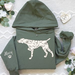 May include: An olive green hoodie featuring a floral dog silhouette. The sleeve displays the name "Macy" with a paw print. The dog graphic is filled with a white floral pattern. A silver necklace is visible.
