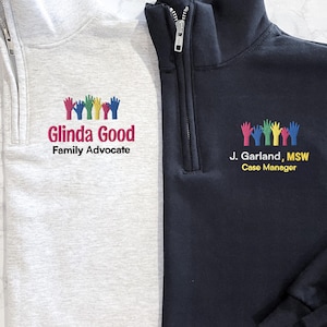 May include: Two quarter-zip pullovers, one light gray and one navy blue. Both feature embroidered colorful hand designs and text. The light gray pullover reads "Glinda Good, Family Advocate." The navy blue pullover reads "J. Garland, MSW, Case Manager."