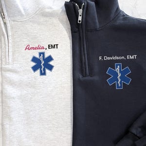 Könnte beinhalten: Zwei Pullover mit Viertelreißverschluss, einer hellgrau und einer marineblau. Jeder hat ein gesticktes blaues Star of Life-Emblem und den Text "Amelia, EMT" oder "F. Davidson, EMT". Die Pullover haben einen hohen Kragen und einen silbernen Reißverschluss.