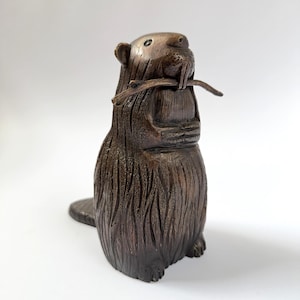 May include: A brown, carved wooden beaver figurine. The beaver is standing upright, holding a small branch in its mouth. The figurine has detailed fur texture and a flat tail. The beaver is a decorative item.