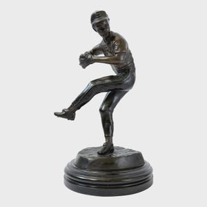 Könnte beinhalten: Eine Bronzeskulptur eines Baseball-Pitchers in Aktion. Die Figur, mit Kappe und Uniform, wird in der Mitte des Wurfs dargestellt, montiert auf einem kreisförmigen Sockel. Die Skulptur hat eine dunkle Patina mit detaillierter Darstellung.