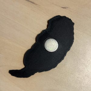 May include: A black embroidered patch with a textured fabric surface and a circular silver-colored metal magnet attached to the center of the back. The patch has an irregular, organic shape with a pointed tail-like extension at the bottom.