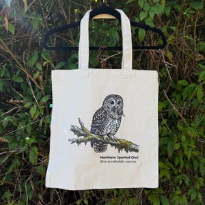 Pu&ograve; includere: Una borsa tote in tela color naturale con un'illustrazione dettagliata di un allocco macchiato del nord appollaiato su un ramo muschioso. Il gufo &egrave; raffigurato in toni di nero, bianco e grigio, mentre tiene una piccola preda. Testo: Northern Spotted Owl.
