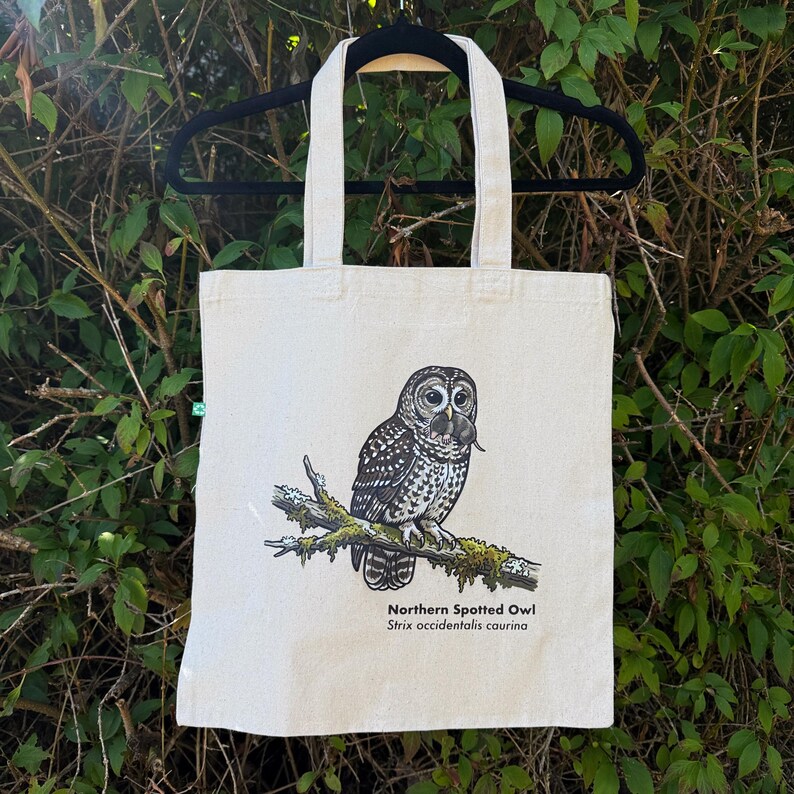 Pu&ograve; includere: Una borsa tote in tela color naturale con un'illustrazione dettagliata di un allocco macchiato del nord appollaiato su un ramo muschioso. Il gufo &egrave; raffigurato in toni di nero, bianco e grigio, mentre tiene una piccola preda. Testo: Northern Spotted Owl.