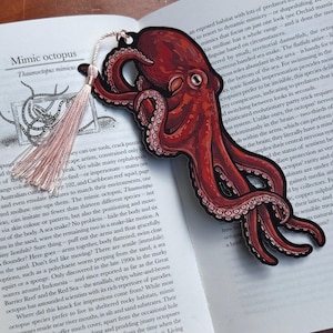 May include: A red and black octopus bookmark with a tassel, resting on an open book. The bookmark features a detailed illustration of an octopus with its tentacles. The book's text includes the words "Mimic octopus".