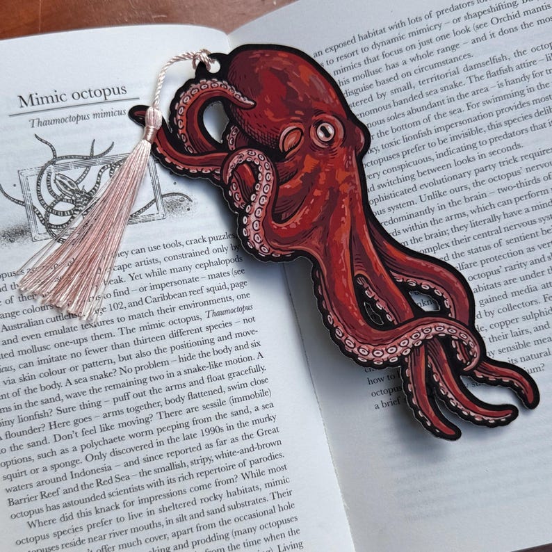 May include: A red and black octopus bookmark with a tassel, resting on an open book. The bookmark features a detailed illustration of an octopus with its tentacles. The book's text includes the words "Mimic octopus".