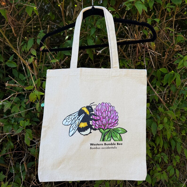 K&ouml;nnte beinhalten: Eine beigefarbene Tragetasche aus Segeltuch mit einer grafischen Illustration einer Westlichen Hummel, Bombus occidentalis, die auf einer rosa Kleepflanze sitzt. Das Design enth&auml;lt den in schwarzer Schrift gedruckten Text Western Bumble Bee und Bombus occidentalis.