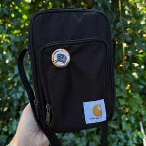 May include: A black rectangular crossbody bag with a front zippered pocket, a black shoulder strap, and a white square patch featuring the Carhartt logo. A circular pin with a raccoon illustration and the text JIMOTHY FOREVER is attached.