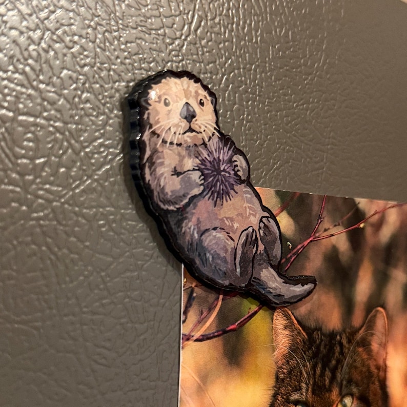 May include: A die-cut refrigerator magnet featuring a detailed illustration of a brown sea otter floating on its back. The otter has a light-colored face and dark whiskers, and it is holding a spiky purple sea urchin against its chest.