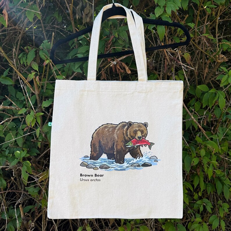 May include: A beige canvas tote bag featuring a graphic illustration of a brown bear standing in blue water with a salmon in its mouth. Below the illustration, the text reads Brown Bear and Ursus arctos in a simple black sans-serif font.