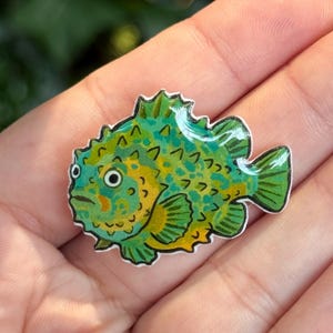 Puede incluir: Un pin plano y brillante con forma de pez espinoso en una paleta de colores vibrantes de verde lima, amarillo y azul cerceta. El pez presenta un cuerpo redondeado, aletas prominentes y ojos grandes y separados, todo delineado en negro.