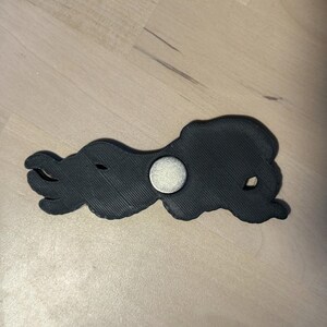 May include: A black, 3D-printed octopus-shaped object with a textured surface and a circular silver-colored metal magnet embedded in the center. The octopus has multiple curved tentacles extending to the left and a rounded head on the right side.
