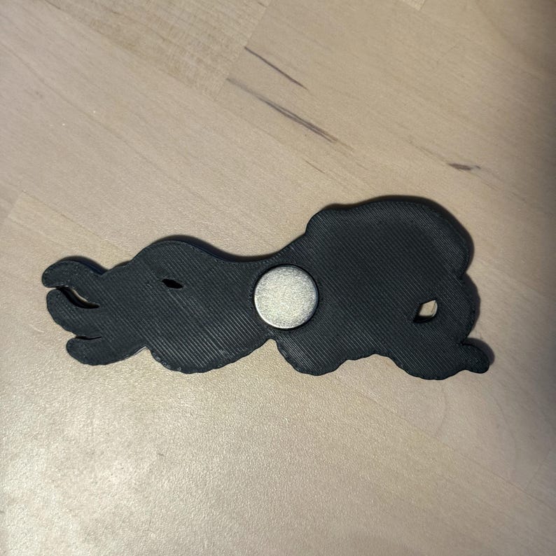May include: A black, 3D-printed octopus-shaped object with a textured surface and a circular silver-colored metal magnet embedded in the center. The octopus has multiple curved tentacles extending to the left and a rounded head on the right side.