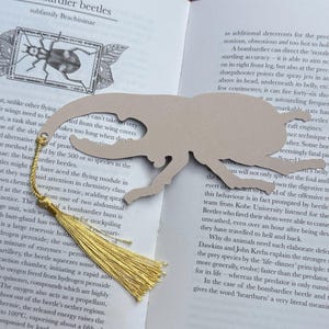 K&ouml;nnte beinhalten: Lesezeichen in K&auml;ferform aus beigem Karton mit goldener Quaste. Das Lesezeichen liegt auf einer Buchseite mit Text und einer K&auml;ferillustration. Ein einzigartiges Accessoire f&uuml;r Leser.