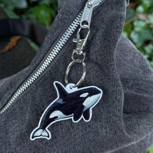 Puede incluir: Un llavero de orca en blanco y negro con un mosquetón y un anillo de metal plateado. El llavero está sujeto a una bolsa de mezclilla gris oscuro con una cremallera plateada. La orca tiene marcas blancas.