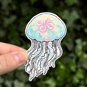 Pu&ograve; includere: Un adesivo fustellato raffigurante un'illustrazione stilizzata di una medusa con una campana azzurra traslucida e una prominente forma rosa a quattro petali al centro. La medusa ha lunghi tentacoli ondulati e fluidi nei toni del blu e del rosa.