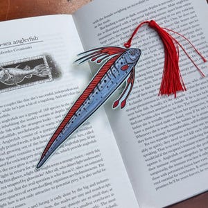 Peut inclure: Marque-page en forme de poisson-p&ecirc;cheur avec un pompon rouge. Le marque-page pr&eacute;sente une illustration d&eacute;taill&eacute;e d'une baudroie bleue et rouge, cr&eacute;ature des abysses, avec un long corps et des nageoires. Le marque-page est pos&eacute; sur un livre ouvert.