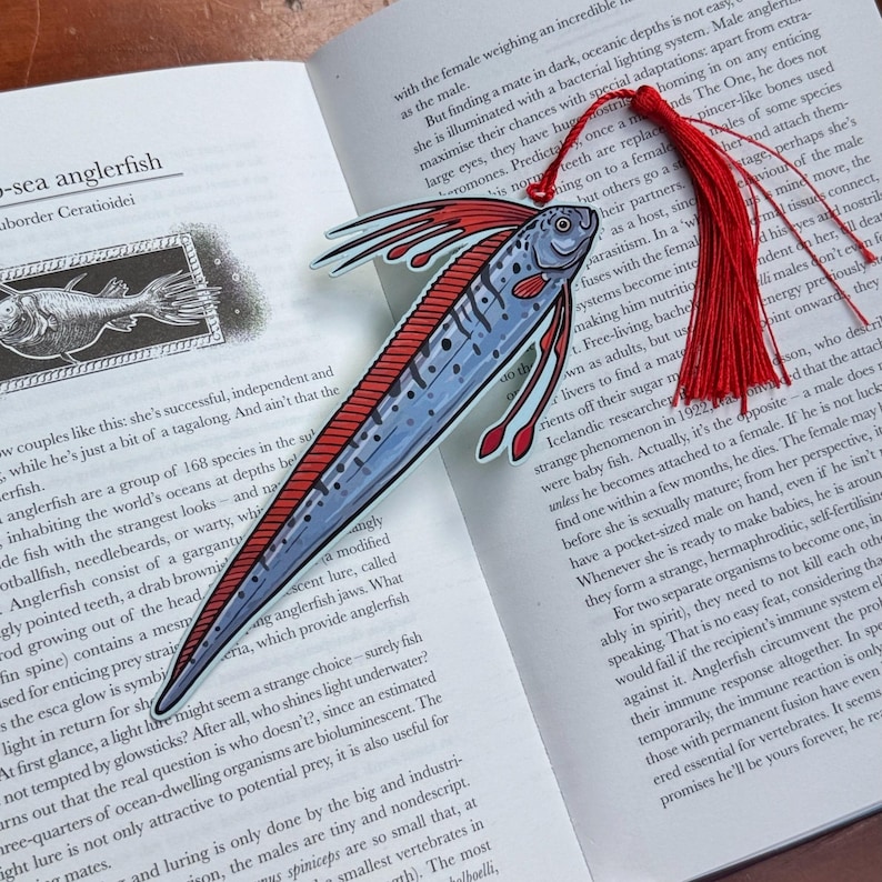 Peut inclure: Marque-page en forme de poisson-p&ecirc;cheur avec un pompon rouge. Le marque-page pr&eacute;sente une illustration d&eacute;taill&eacute;e d'une baudroie bleue et rouge, cr&eacute;ature des abysses, avec un long corps et des nageoires. Le marque-page est pos&eacute; sur un livre ouvert.