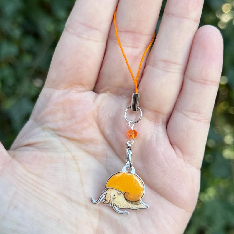 May include: A silver charm with an orange snail design, attached to an orange cord. The snail has a yellow shell and a cream-colored body. The charm is connected to the cord with a silver clasp and a small orange bead.