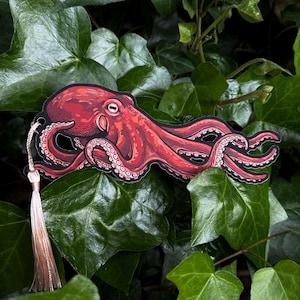 May include: A red octopus bookmark with a cream-colored tassel. The octopus illustration has detailed shading and suction cups on its tentacles. The bookmark is set against a backdrop of green leaves, creating a natural contrast.