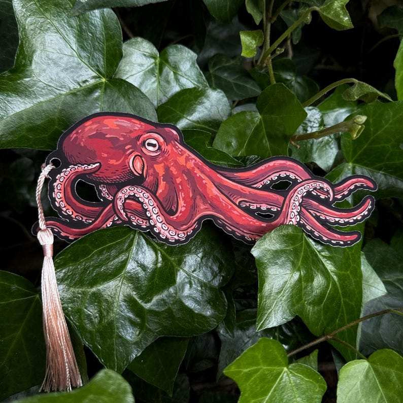 May include: A red octopus bookmark with a cream-colored tassel. The octopus illustration has detailed shading and suction cups on its tentacles. The bookmark is set against a backdrop of green leaves, creating a natural contrast.