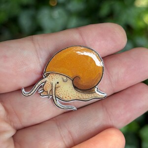 Puede incluir: Un pin plano y brillante con una ilustraci&oacute;n estilizada de un caracol con una concha redondeada de color naranja vibrante y un cuerpo de color beige claro cubierto de peque&ntilde;as motas naranjas. El caracol tiene dos tent&aacute;culos largos y finos.