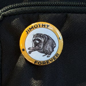 May include: A circular pinback button featuring a black and white illustration of a raccoon centered on a light gray background. The button has a yellow border with the black text JIMOTHY at the top and FOREVER at the bottom, separated by stars.