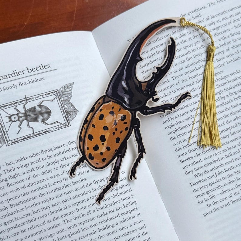 K&ouml;nnte beinhalten: Ein detaillierter Lesezeichen-Entwurf mit einem schwarz-orangen Bombardierk&auml;fer. Das Lesezeichen hat einen goldenen Quastenanh&auml;nger und liegt auf einem offenen Buch.
