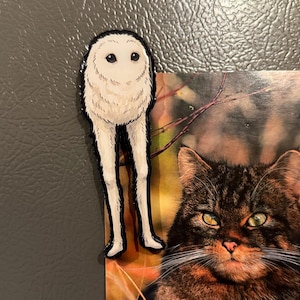May include: A flat, die-cut magnet featuring a stylized, round, white, fluffy creature with two long, slender legs and a small, dark face. The creature has a soft, textured appearance with dark outlines and is positioned next to a cat photograph.