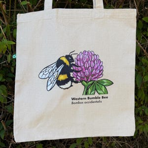 K&ouml;nnte beinhalten: Eine naturfarbene Stofftasche mit einer grafischen Illustration einer Westlichen Hummel, Bombus occidentalis, die auf einer rosa Klee-Bl&uuml;te sitzt. Das Design enth&auml;lt den schwarzen Schriftzug Western Bumble Bee und Bombus occidentalis.