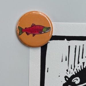 May include: A round orange button featuring a red fish with green and black details. The fish is swimming to the left. The button is on a white surface with a black and white design in the background.
