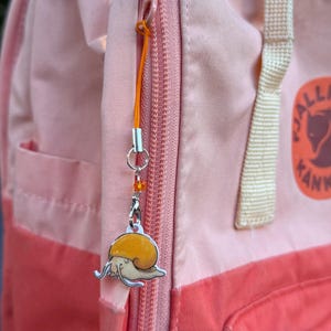 May include: A snail charm with an orange shell and a silver outline hangs from an orange cord attached to a pink backpack zipper. The charm is detailed with a cream-colored body and a silver metal finish.