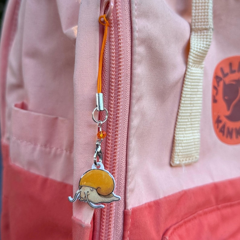 May include: A snail charm with an orange shell and a silver outline hangs from an orange cord attached to a pink backpack zipper. The charm is detailed with a cream-colored body and a silver metal finish.