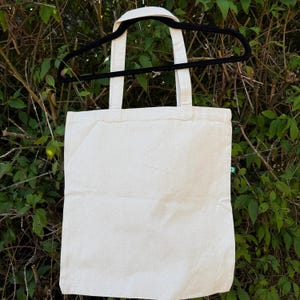 Pu&ograve; includere: Una borsa tote in tela color bianco sporco con due manici in tessuto abbinati, appesa a una gruccia di plastica nera. La borsa presenta una piccola etichetta rettangolare verde cucita sulla cucitura laterale, con un'icona di riciclo bianca.