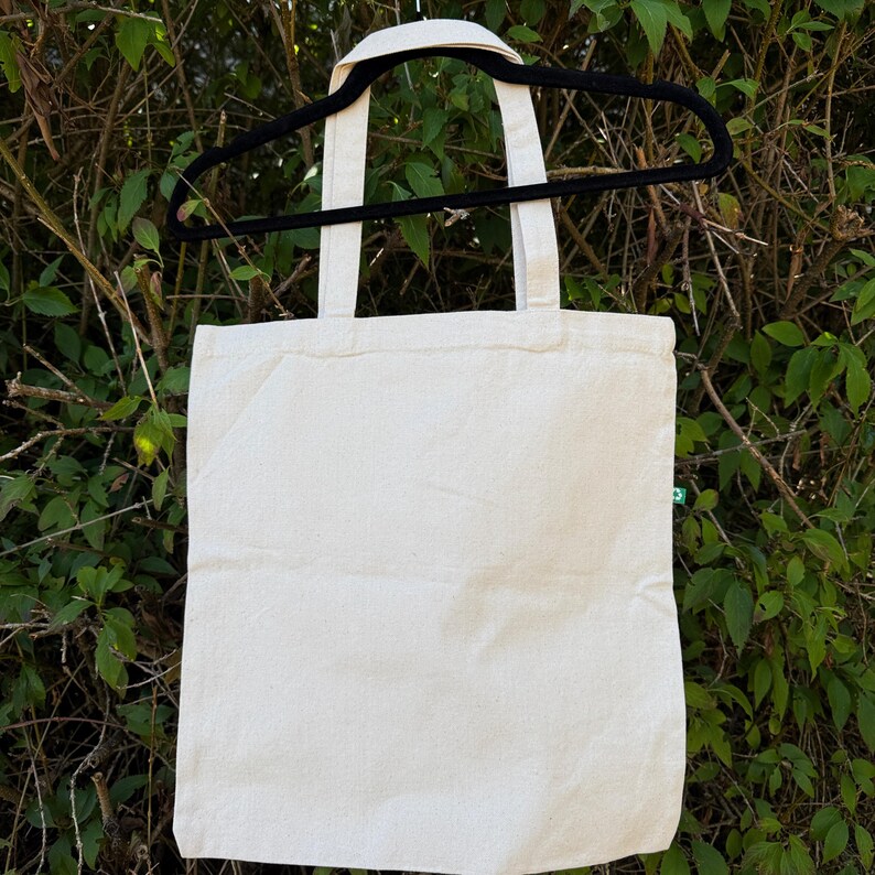 Pu&ograve; includere: Una borsa tote in tela color bianco sporco con due manici in tessuto abbinati, appesa a una gruccia di plastica nera. La borsa presenta una piccola etichetta rettangolare verde cucita sulla cucitura laterale, con un'icona di riciclo bianca.