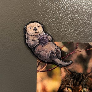 May include: A die-cut magnet featuring a brown sea otter with a light-colored face, holding a purple sea urchin against its chest. The otter has a dark nose, whiskers, and a textured fur appearance, depicted in a realistic illustrative style.