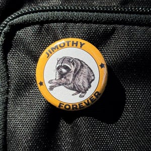 May include: A circular pin-back button featuring a central illustration of a gray and black raccoon on a white background. The border is yellow with the black text JIMOTHY at the top and FOREVER at the bottom, accented by two small black stars.