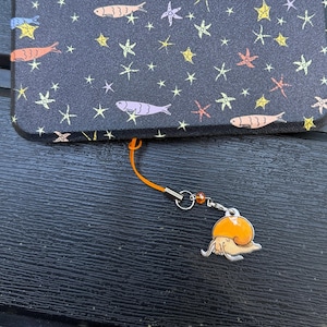 May include: A black phone case with a pattern of colorful fish and stars. An orange lanyard is attached to the case, with a silver charm of an orange snail.