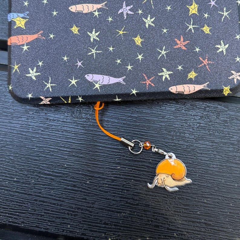 May include: A black phone case with a pattern of colorful fish and stars. An orange lanyard is attached to the case, with a silver charm of an orange snail.