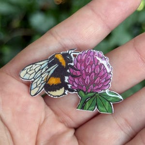 Puede incluir: Un pin plano que presenta una ilustraci&oacute;n detallada de un abejorro peludo con rayas negras y amarillas y alas transl&uacute;cidas, colocado junto a una flor de tr&eacute;bol de color p&uacute;rpura vibrante con hojas verdes, con un acabado brillante y claro.